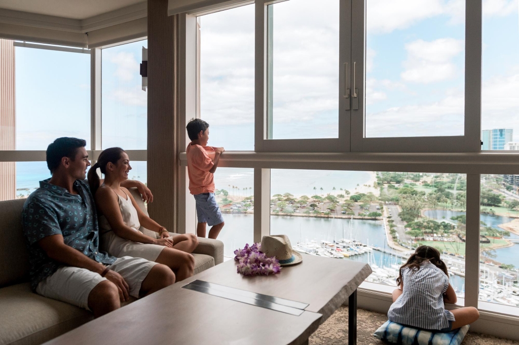 A family of four - father, mother, son and daughter - enjoying the ocean views from the floor-to-ceiling windows at Prince Waikiki.