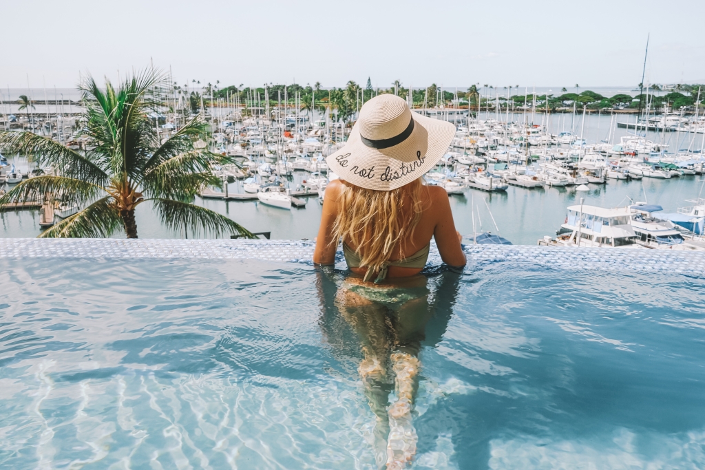 A woman in the infinity pool with a straw hat on overlooking the ocean yacht harbor view at Prince Waikiki.